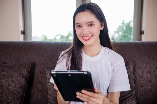 Asian young woman happy cheerful cute beautiful is playing digital pad tablet computer and sitting on the sofa at home, smiling lady using app reading electronic book relax in living room 