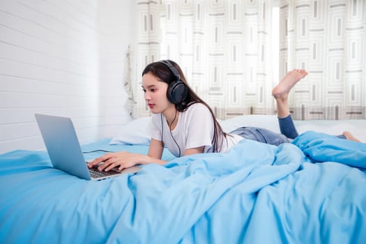 Asian happy young woman wear headphones and listening music song and watching website for shopping online from laptop  on bed at home