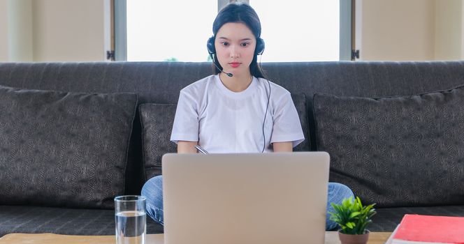 Asian happy woman student wear headphones study online watching webinar podcast on laptop listening learning education course conference calling and write notes look at laptop sit on sofa at home
