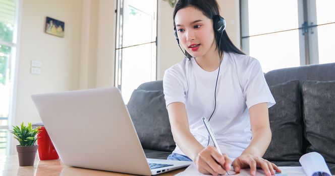 Asian happy woman student wear headphones study online watching webinar podcast on laptop listening learning education course conference calling and write notes look at laptop sit on sofa at home