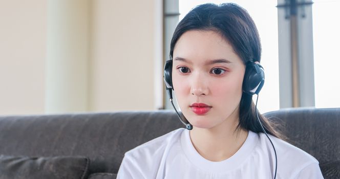 Asian happy woman student wear headphones study online watching webinar podcast on laptop listening learning education course conference calling and write notes look at laptop sit on sofa at home