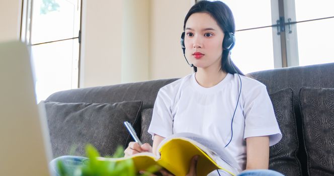 Asian happy woman student wear headphones study online watching webinar podcast on laptop listening learning education course conference calling and write notes look at laptop sit on sofa at home