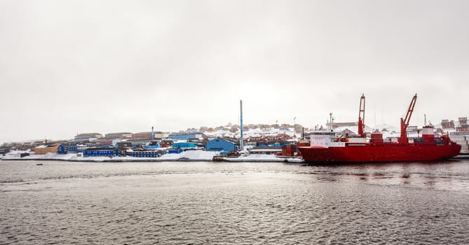 Big red cargo ship under the snowfall in the port of Aasiaat, with village in the background, Greenland