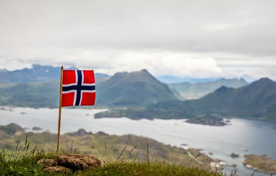 Norwegian national flag in the wind on the top of Nonstinden peak with fjord in the background, Ballstad, Vestvagoy Municipality, Nordland county, Norway