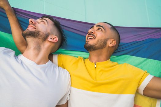Portrait of young gay couple embracing and showing their love with rainbow flag in the street. LGBT and love concept.