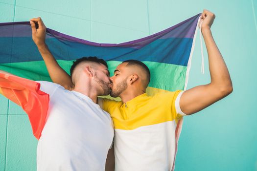 Portrait of young gay couple embracing and showing their love with rainbow flag in the street. LGBT and love concept.
