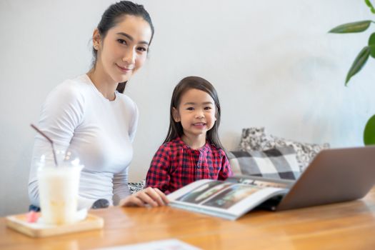 Asian mothers are teaching their daughters to read a book and use notebooks and technology for online learning during school holidays at home. Educational concepts and activities of the family