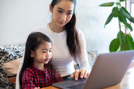Asian mothers are teaching their daughters to read a book and use notebooks and technology for online learning during school holidays at home. Educational concepts and activities of the family