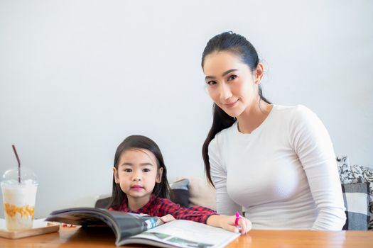An Asian mother is teaching her daughter to read a book during the semester break on the living table and having cold milk on the table at home. Educational concepts and activities of the family