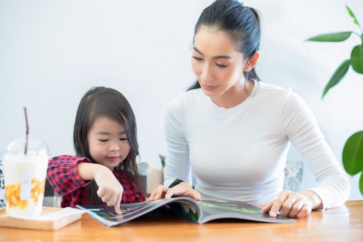 An Asian mother is teaching her daughter to read a book during the semester break on the living table and having cold milk on the table at home. Educational concepts and activities of the family