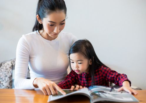 An Asian mother is teaching her daughter to read a book during the semester break on the living table and having cold milk on the table at home. Educational concepts and activities of the family