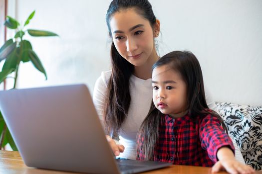 Asian mothers are teaching their daughters to read a book and use notebooks and technology for online learning during school holidays at home. Educational concepts and activities of the family