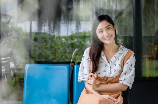Cute Asian woman sits on the sofa or chair and looks at the camera and smiles happy in portraits at home during relax time.