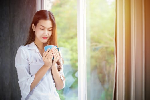 An Asian woman stood holding a coffee cup and opened the curtain on the window after waking up in the morning of the day at home