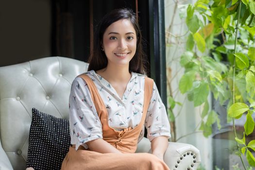 Cute Asian woman sits on the sofa or chair and looks at the camera and smiles happy in portraits at home during relax time.