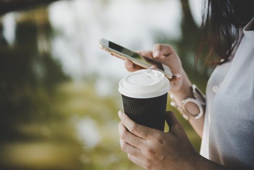 Young woman holding disposable coffee cup while text messaging through smartphone at nature outdoors.