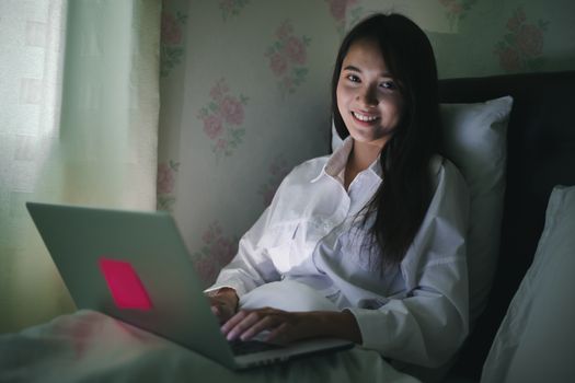 Asian woman using a notebook to work on night at the bed.She smiles and enjoys working at home.