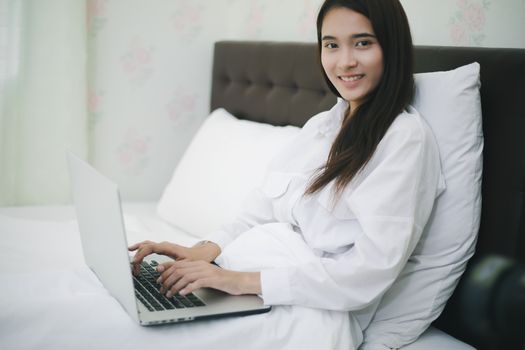 Asian woman using a notebook to work on the bed.She smiles and enjoys working at home.