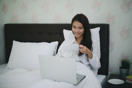 Asian woman is drinking a cup of coffee and using a notebook to work on the bed.She smiles and enjoys working at home.