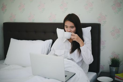 Asian woman is drinking a cup of coffee and using a notebook to work on the bed.She smiles and enjoys working at home.