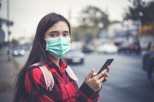 Asian Young woman using smart phone in the city wearing face mask because of air pollution, particulates and for protection flu virus, influenza, coronavirus
