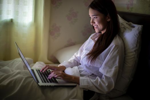 Asian woman using a notebook to work on night at the bed.She smiles and enjoys working at home.