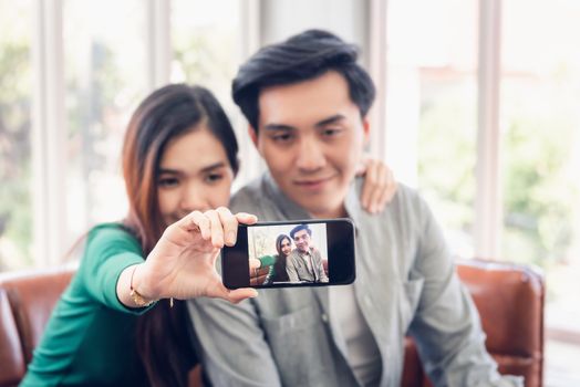 Portrait of Young Couple Love Capturing Selfie on Mobile Phone at Their Home, Attractive Asian Couple Making Selfie and Smile While Resting on a Couch. Relaxing and Enjoyment Time
