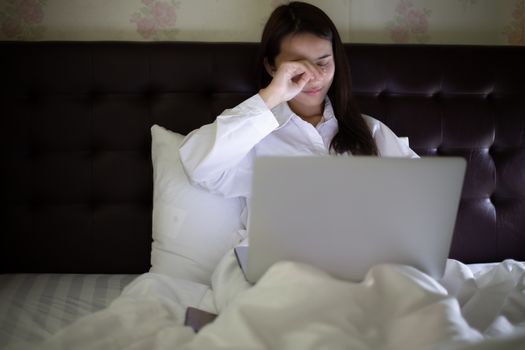 Asian woman using a notebook to work at night in bed and she yawns and sleepy.