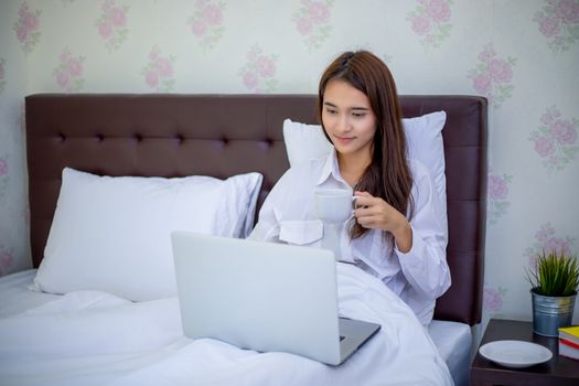 Asian woman is drinking a cup of coffee and using a notebook to work on the bed.She smiles and enjoys working at home.