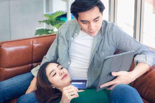 Young Couple Love Having Enjoyment While Online Shopping on Electronic Tablet in Living Room, Portrait of Asian Couple Relaxing on a Couching During Shopping Online Togetherness. Relaxation/ Lifestyle