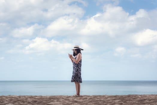 Portrait of Asian Woman is Using Mobile Phone Against Blue Sky and Sand Beach, Beautiful Woman is Having Fun and Enjoying With Her Phone While Summer Vacation on The Beach. Relaxation Lifestyles