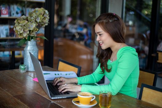 Asian businesswomen smiling and using notebook for analysis documents and graph financial diagram working and she is drinking coffee at coffee shop