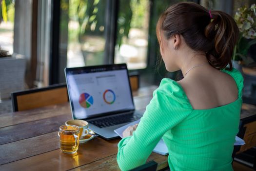 Asian businesswomen smiling and using notebook for analysis documents and graph financial diagram working and she is drinking coffee at coffee shop