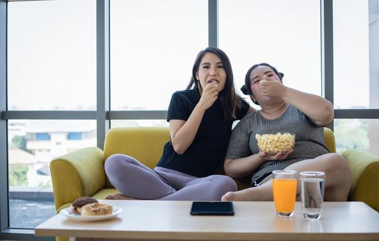 Overweight woman and asian girl enjoy eating food on sofa at home