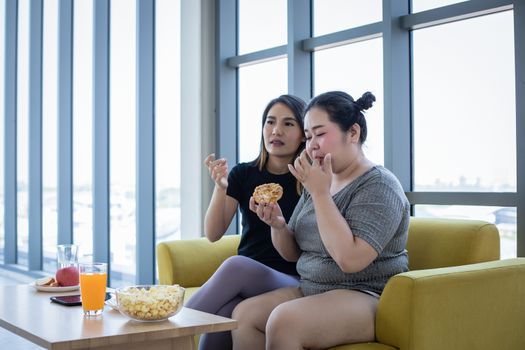 Overweight woman and asian girl enjoy eating food on sofa at home