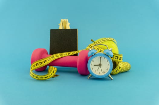 Starting healthy lifestyle concept. Still life with pink dumbbell, measuring tape, apple, chalkboard and alarm clock on blue background