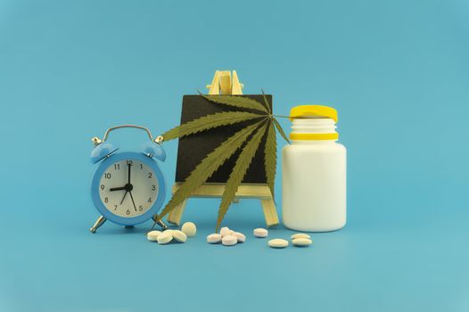 Medicinal marijuana concept with fresh green leaf, pharmaceutical bottle and pills, alarm clock and small chalk board over a blue background with copy space