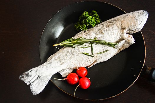 Top view of a trout fish covered in flour topped with rosemary and parsley in an iron skillet and ready to be fried