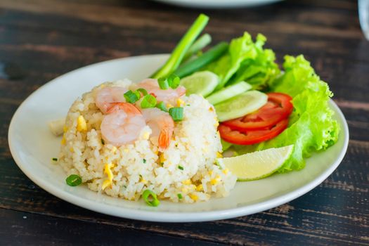 Close up fried rice with shrimp, egg and green onion on white plate serve with blurry cucumber, red tomato, lettuce in cafe and restaurant.