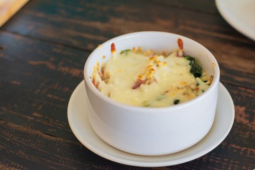Delicious bake spinach with cheese in ceramic white bowl on plate and wooden table in cafe and restaurant.