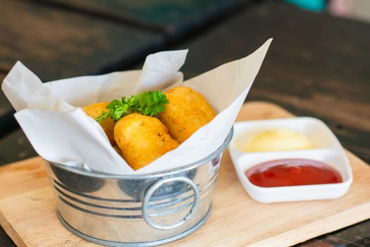 Delicious deep fried chicken nuggets on white paper in metal bucket on wooden tray and rustic table with blurry ketchup and mayonnaise. Unhealthy food concept.