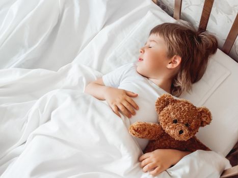 Toddler sleeps in bed with cute teddy bear. Little boy under white blanket with fluffy toy. Plush guard watches out child's sleep. Cozy home.