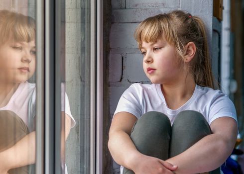 girl of 8 years old with blond hair sits on the windowsill and looks out the window at her reflection