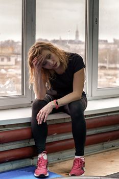 30 years old woman with blond hair sits on the windowsill against the background of a window after a workout in the gym