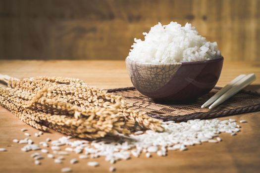 Cooked rice in bowl with raw rice grain and dry rice plant on  wooden table background.