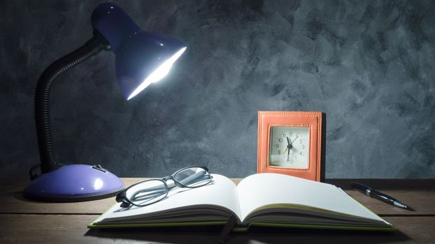 Lamp with notebook,pen,clock and glasses  on  wooden table with vintage wall background.