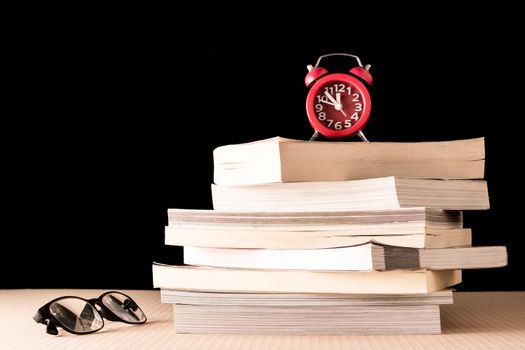 Stack of books with eyeglasses and clock on wooden table, Black background