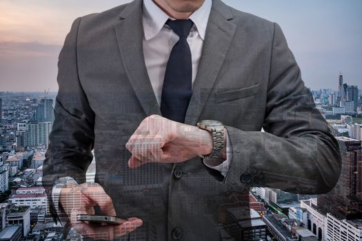 Double exposed of Businessman checking time on his wrist watch and holding mobile phone with cityscape.