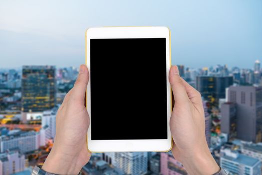Male hands holding a white tablet touch computer gadget with touch blank black screen on blurred city landscape background.