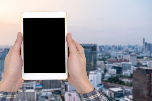 Male hands holding a white tablet touch computer gadget with touch blank black screen on blurred city landscape background.
